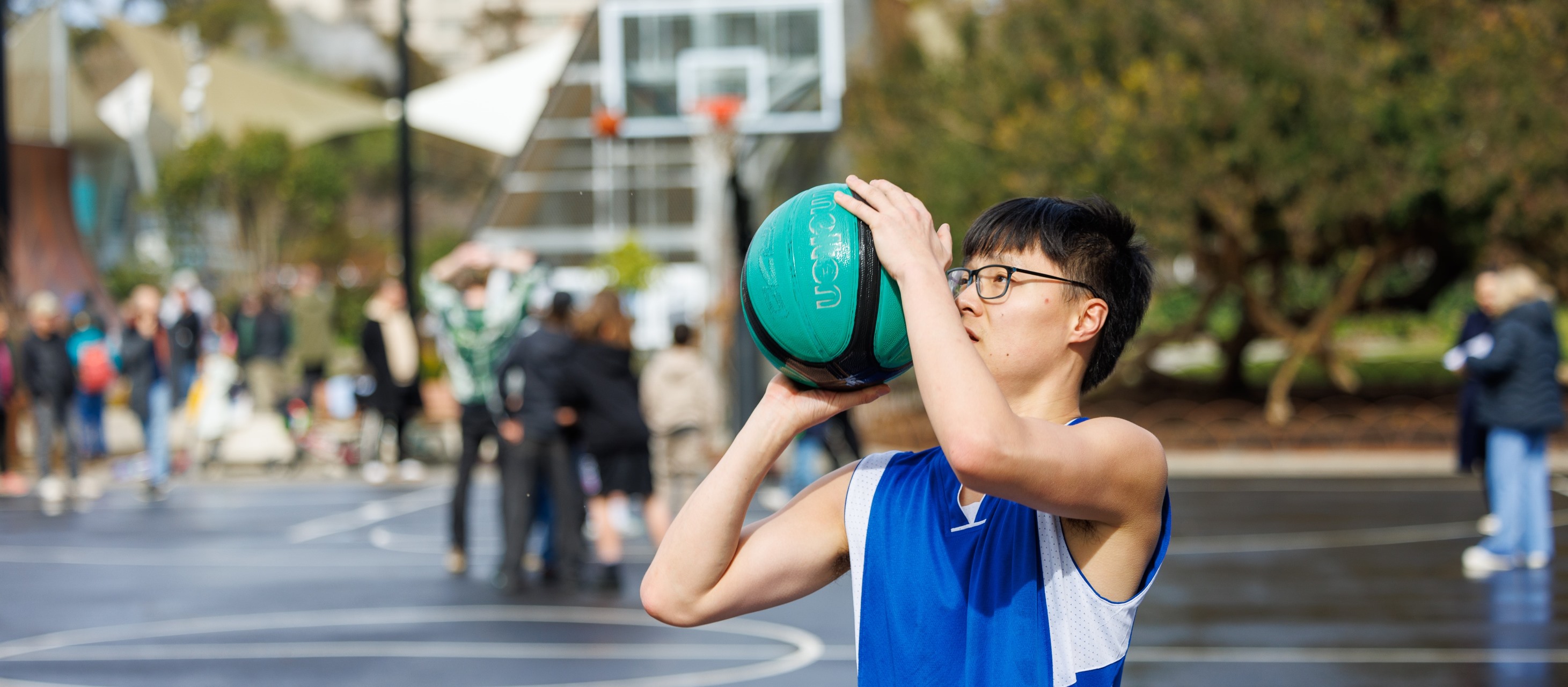 Person in a blue sports top about to shoot a basketball through a hoop. In the background people are enjoying themselves on the court and park.
