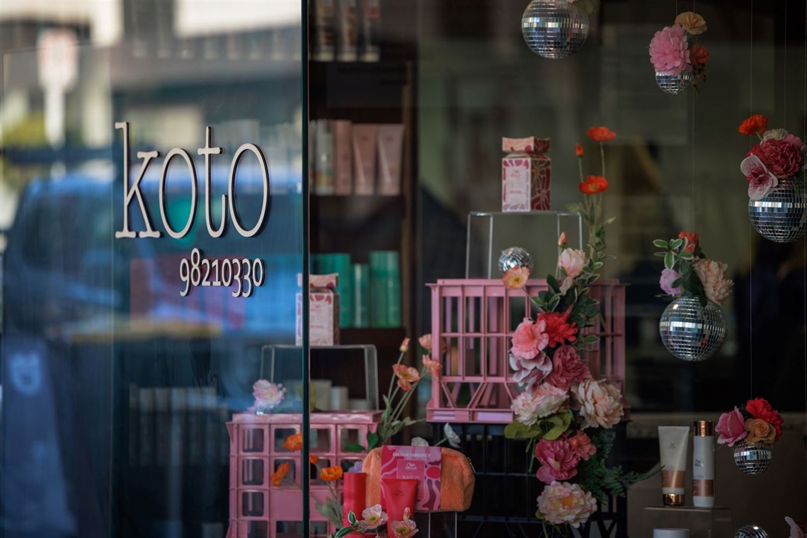 View of a hair salon from the outside. koto 98210330 is on the window. Flowers, disco balls and hair products are inside the salon.