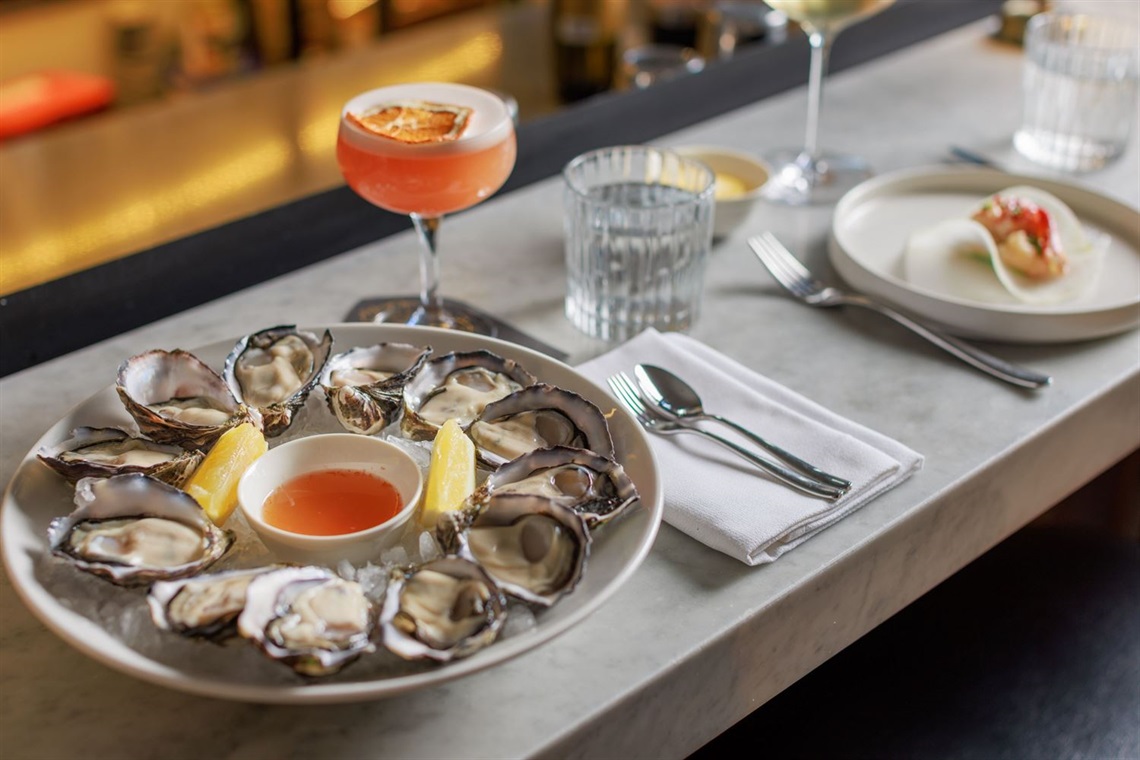 A table with a plate of oysters, a pink cocktail, fine cutlery and other meals.
