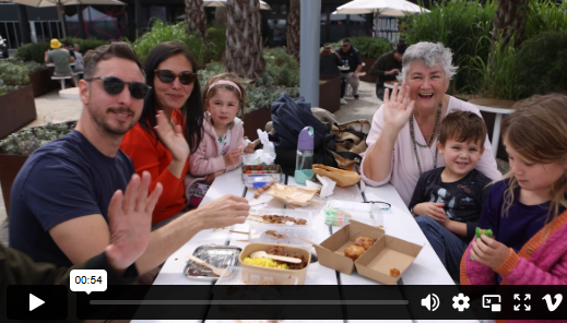 Family sitting at a table enjoying food at the Satay festival. They are smiling and waving at the camera.