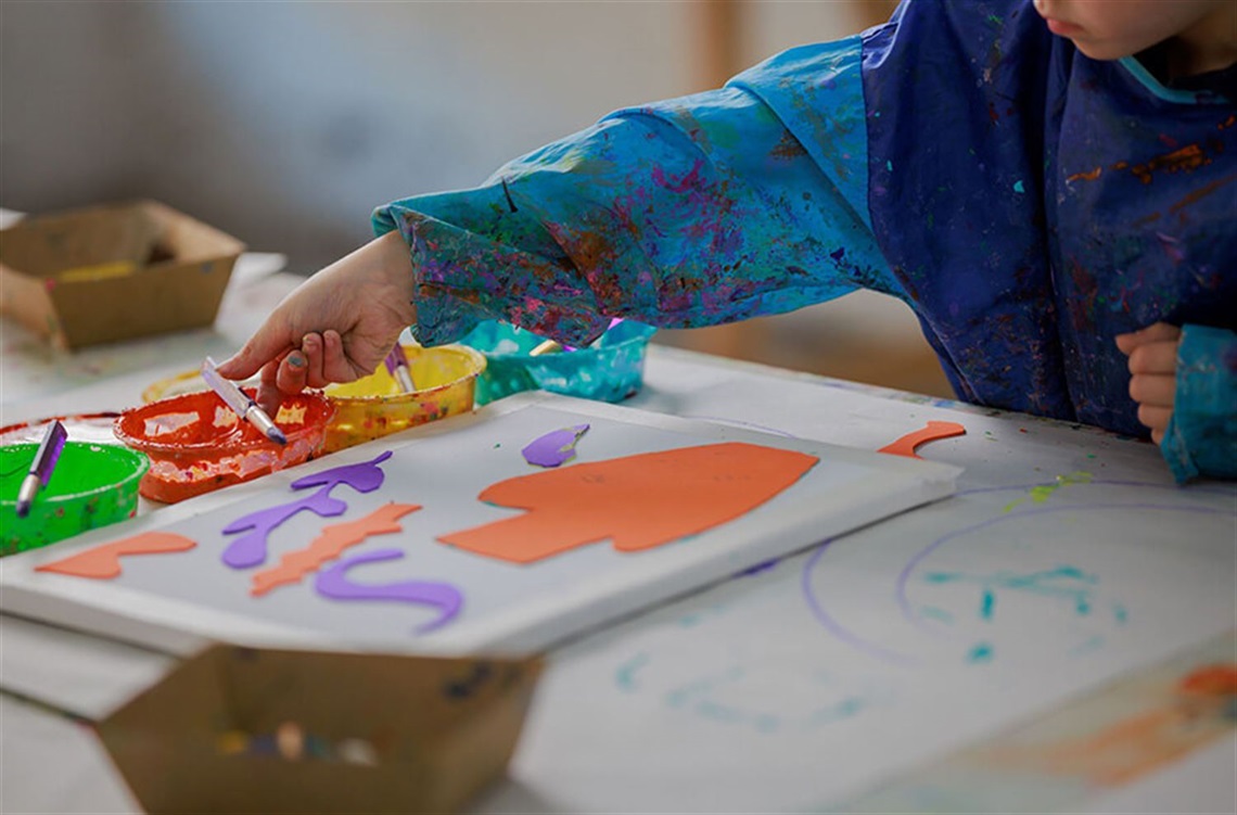 Smiling woman wearing a paint-splattered art smock standing at a table of paints and canvases. She is holding a cut out image of a leaf.