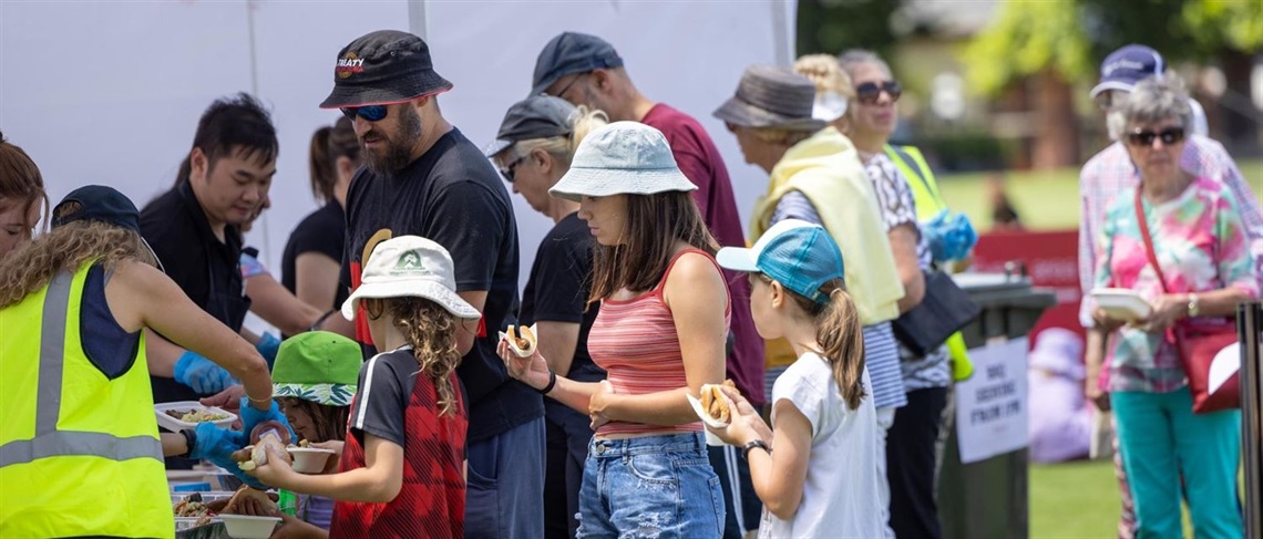 People of various ages standing in line to get a sausage in bread at an outdoor tent in a park.