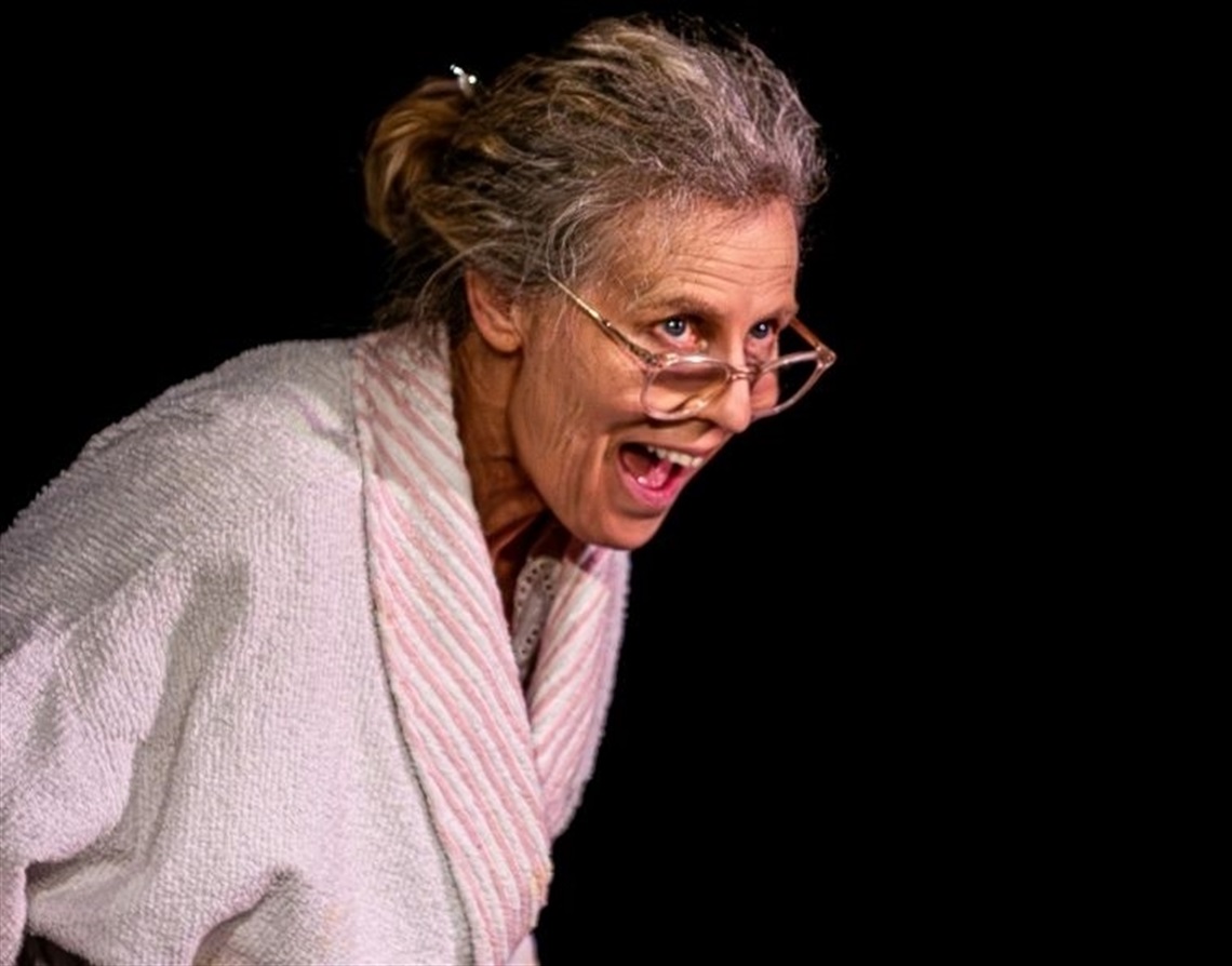 An older woman with fuzzy grey hair in a white dressing gown grinning with an open mouth